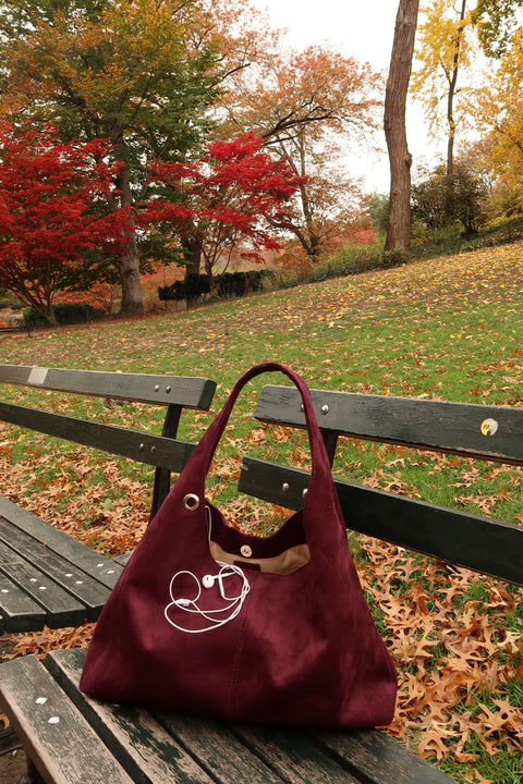 Maroon handbag with a white design on a wooden bench in a park with autumn leaves and trees.